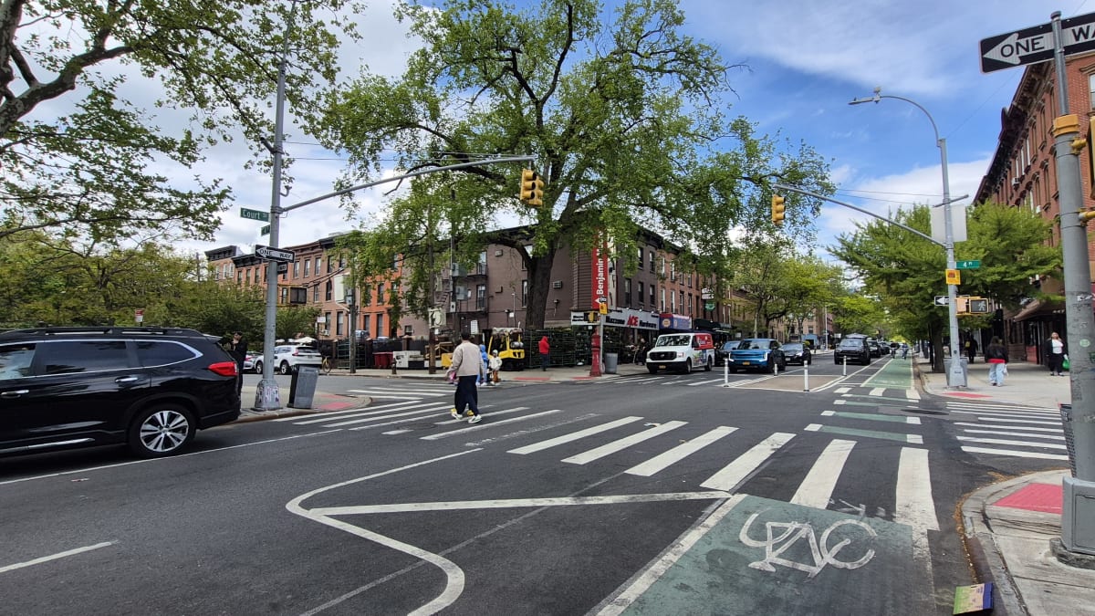 A neighborhood intersection with red-brick walk-ups and storefronts, a bike lane, and a pedestrian crossing