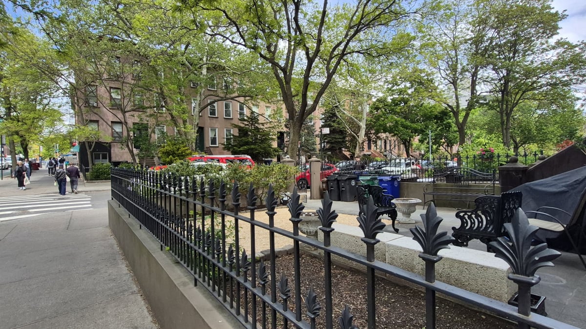 A wrought-iron fence around a leafy garden patio, with brick apartment buildings across the street