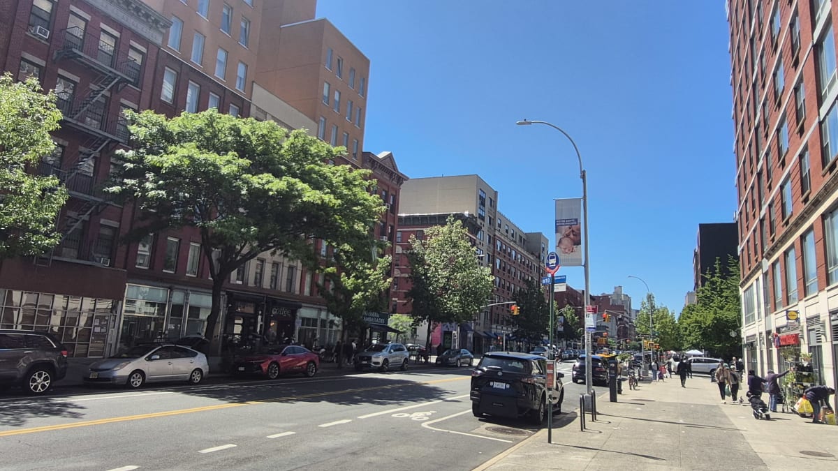 A sunny avenue with red-brick storefront buildings and modern mid-rises, with trees, cars, and pedestrians