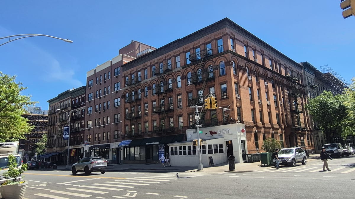 A large ornate red-brick prewar apartment building with storefronts at a sunny street corner