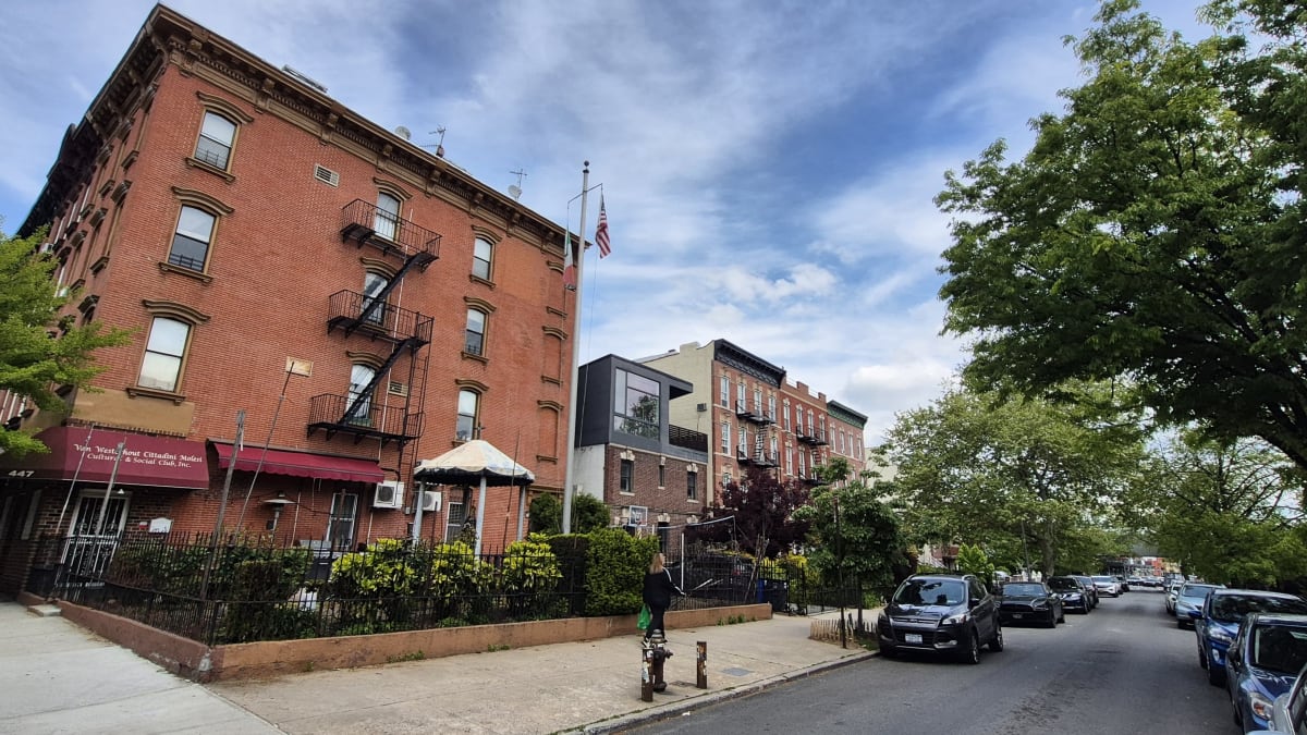 A red-brick prewar walk-up with fire escapes beside a modern townhouse on a tree-lined residential street