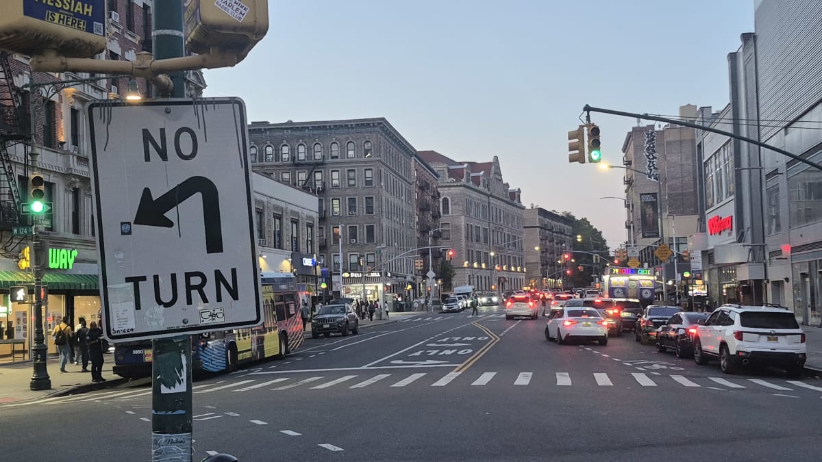 A busy avenue at dusk with a bus, traffic, and prewar apartment buildings above lit storefronts