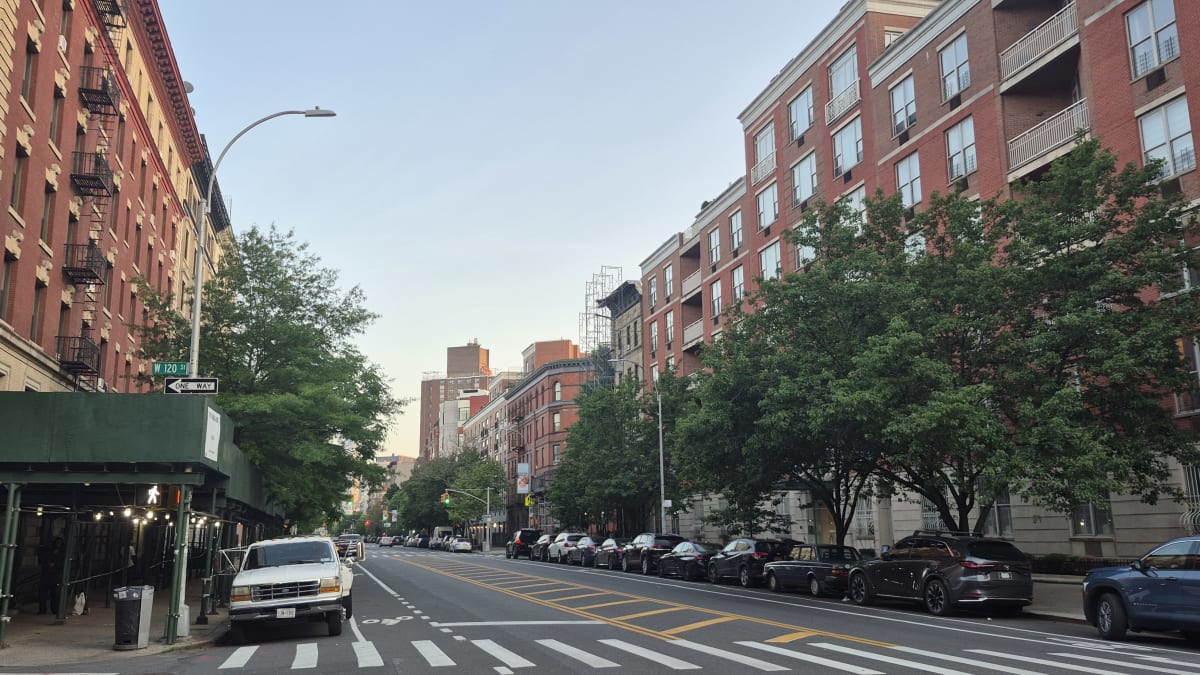 A residential street at dusk lined with red-brick walk-ups and newer balconied buildings, with parked cars