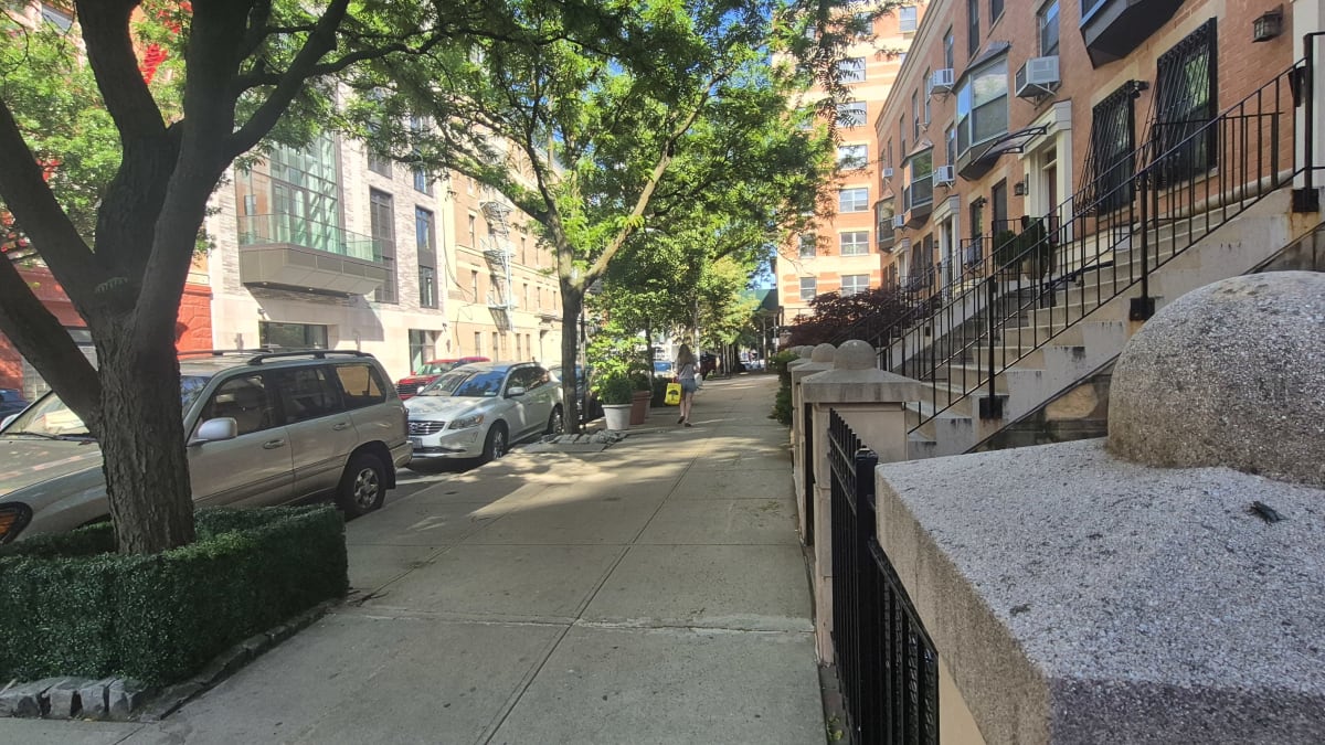 A tree-shaded sidewalk past brick apartment buildings with stoops, with parked cars opposite