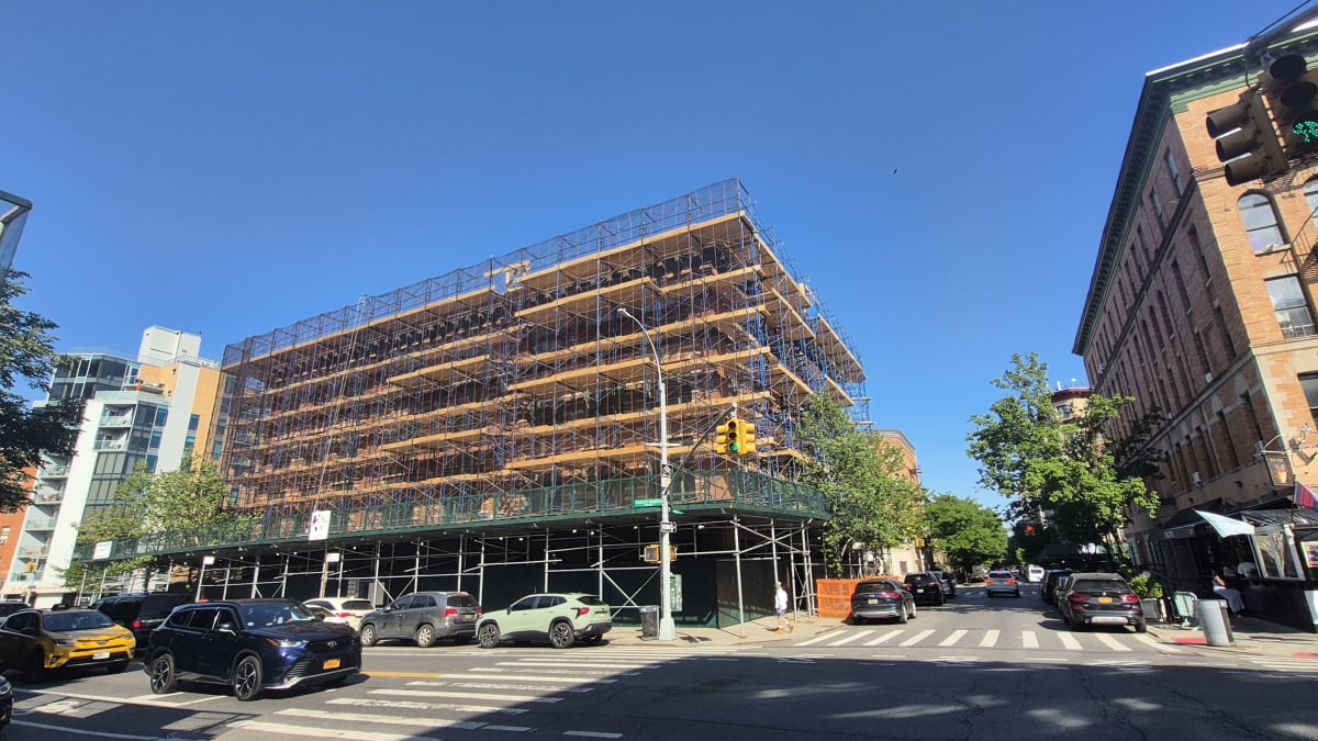 A corner building encased in construction scaffolding at a sunny intersection, with older brick walk-ups nearby