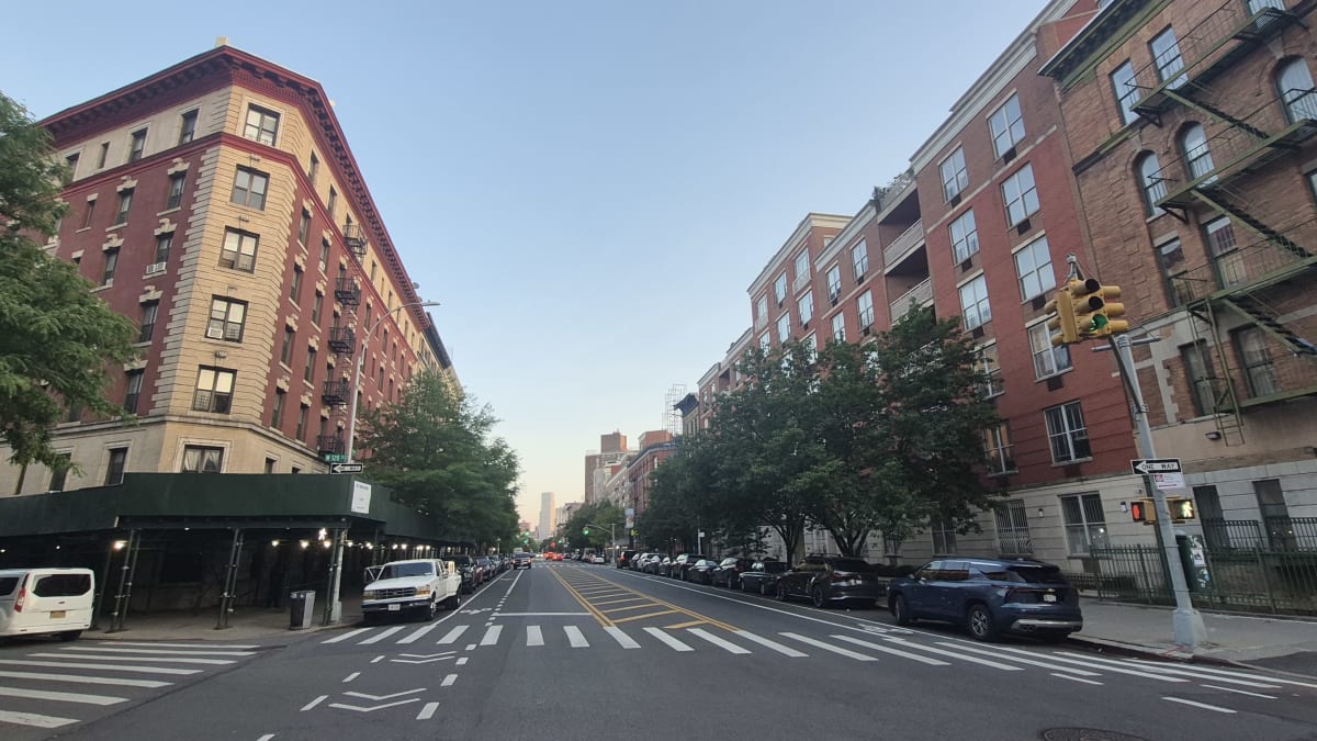 A wide residential avenue at dusk lined with prewar apartment buildings and parked cars