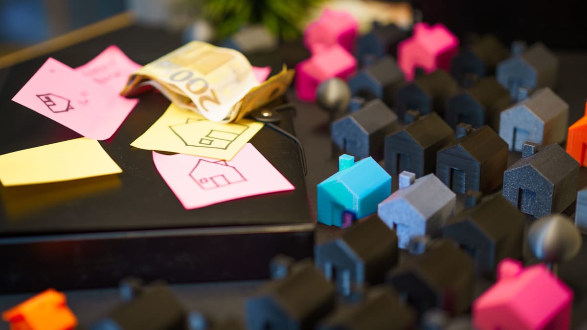 Small model houses and sticky notes sketched with houses beside a folded banknote on a dark surface
