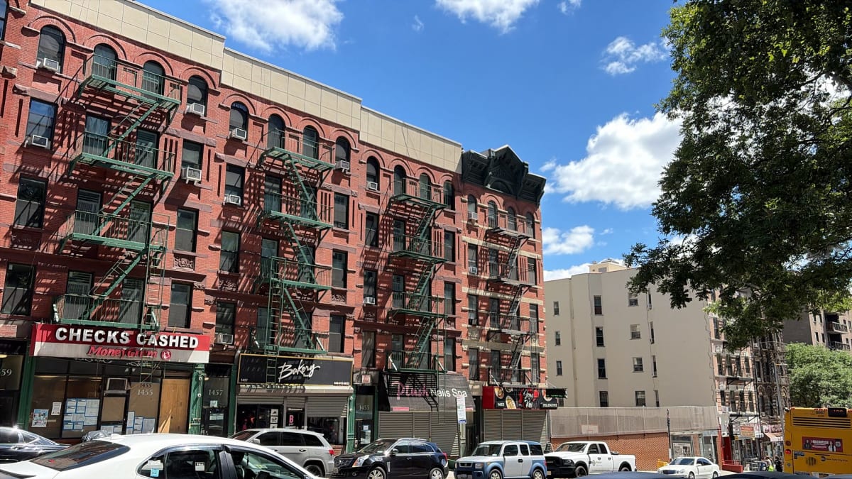 Red-brick prewar walk-ups with green fire escapes above ground-floor storefronts on a sunny street