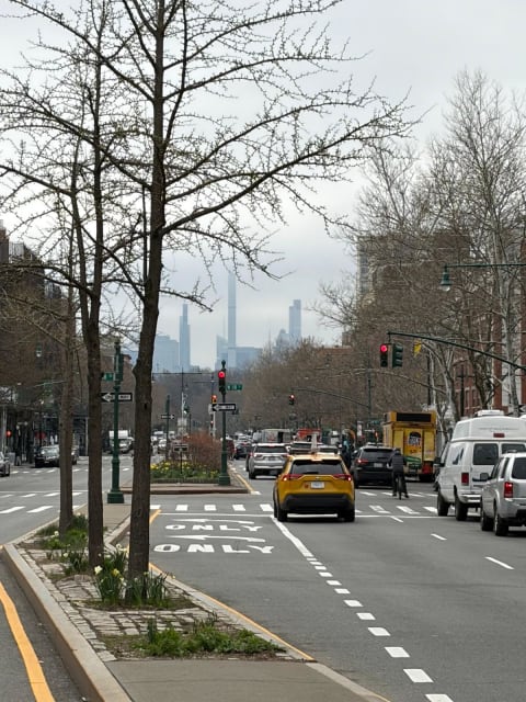 A wide tree-lined boulevard with traffic and the misty Midtown skyline of supertall towers beyond
