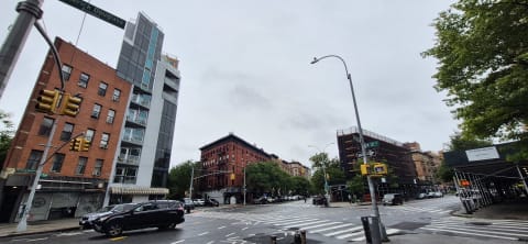 A wide intersection on an overcast day with red-brick walk-ups, a newer balconied building, and traffic