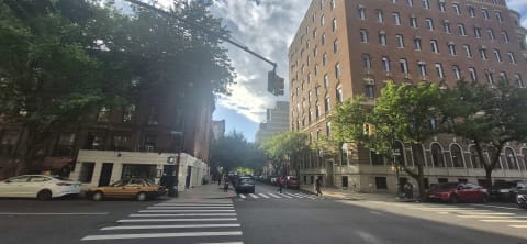 A sunny avenue intersection with large prewar apartment buildings, a taxi, and pedestrians at a crosswalk
