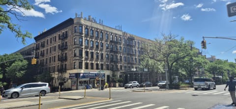A large tan-brick prewar apartment building with fire escapes above a hardware store at a sunny corner