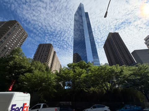 A tall glass skyscraper rising between brick apartment towers above street trees and traffic