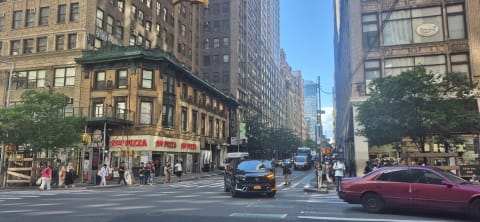 Busy Manhattan avenue intersection with tall prewar buildings, a corner pizza shop, and pedestrians crossing