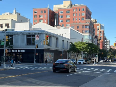 A sunny avenue with a Foot Locker on the corner, mid-rise brick buildings, and crossing traffic