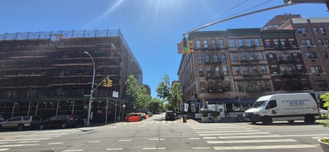A scaffolded building faces red-brick walk-ups with fire escapes across a sunny intersection with crosswalks