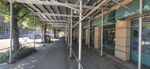 Empty sidewalk beneath a metal construction shed running past storefronts and street trees