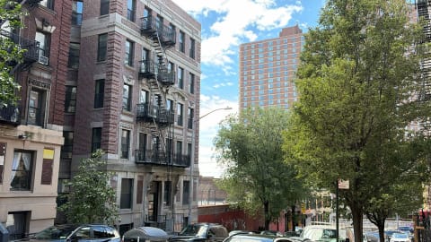 A red-brick prewar walk-up with fire escapes below a taller brick apartment tower, with parked cars