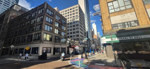 A downtown Manhattan corner of prewar loft buildings with a deli, pedestrians, and a painted plaza