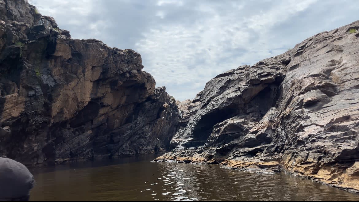 Rugged, towering volcanic rock cliffs form a dramatic narrow gorge over the dark, flowing waters of the Sanaga River under a cloudy sky.