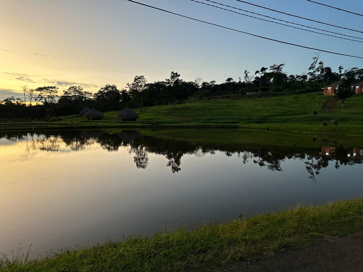 A serene view of the Nkolandom Tourist Center at dusk, featuring traditional thatched-roof huts reflected in a perfectly still lake against a backdrop of dense tropical forest.