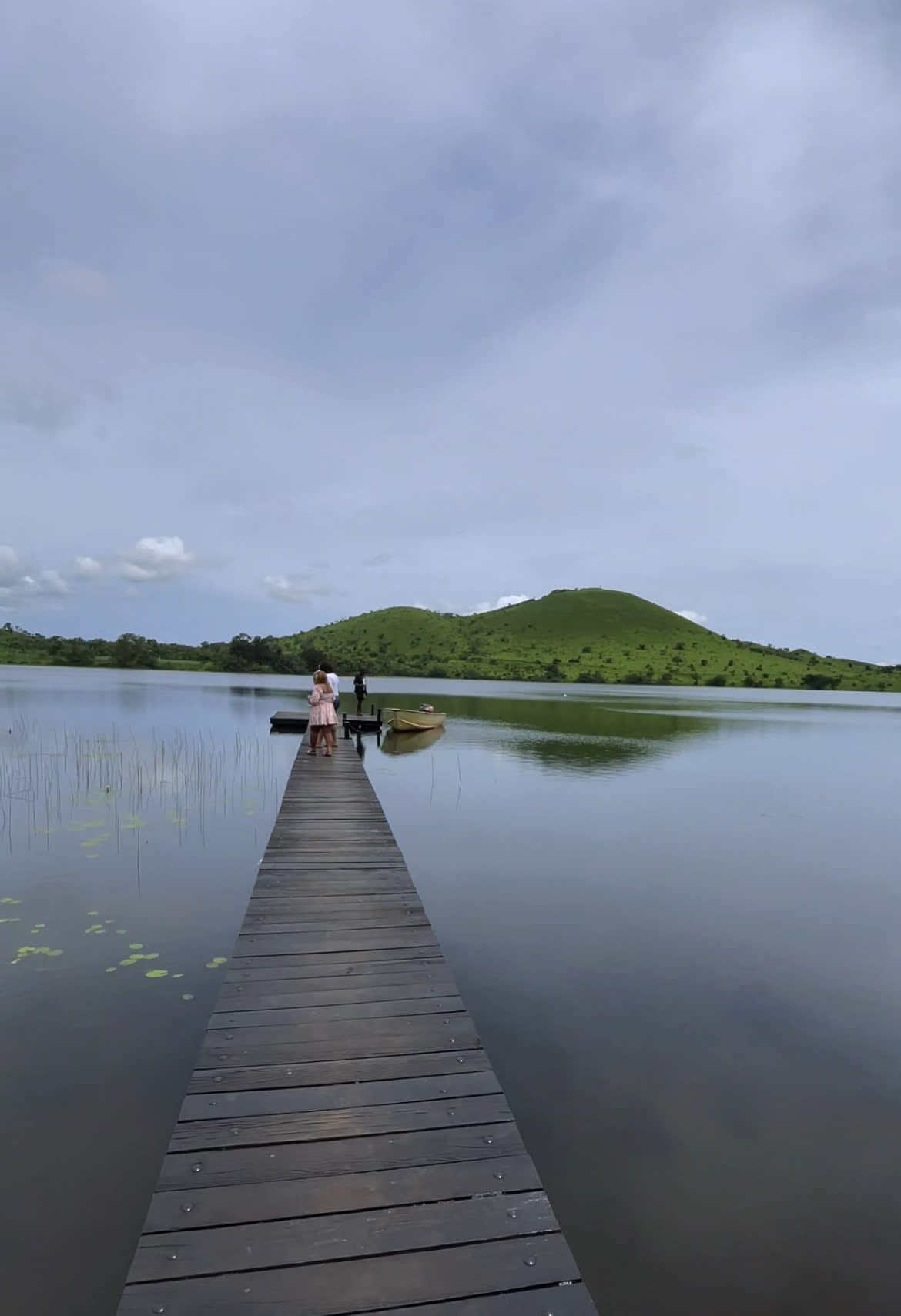 A peaceful wooden pier reaches out across the mirror-like, golden waters of Lake Petpenoun, beautifully framed by distant green mountains under a soft, hazy sky.