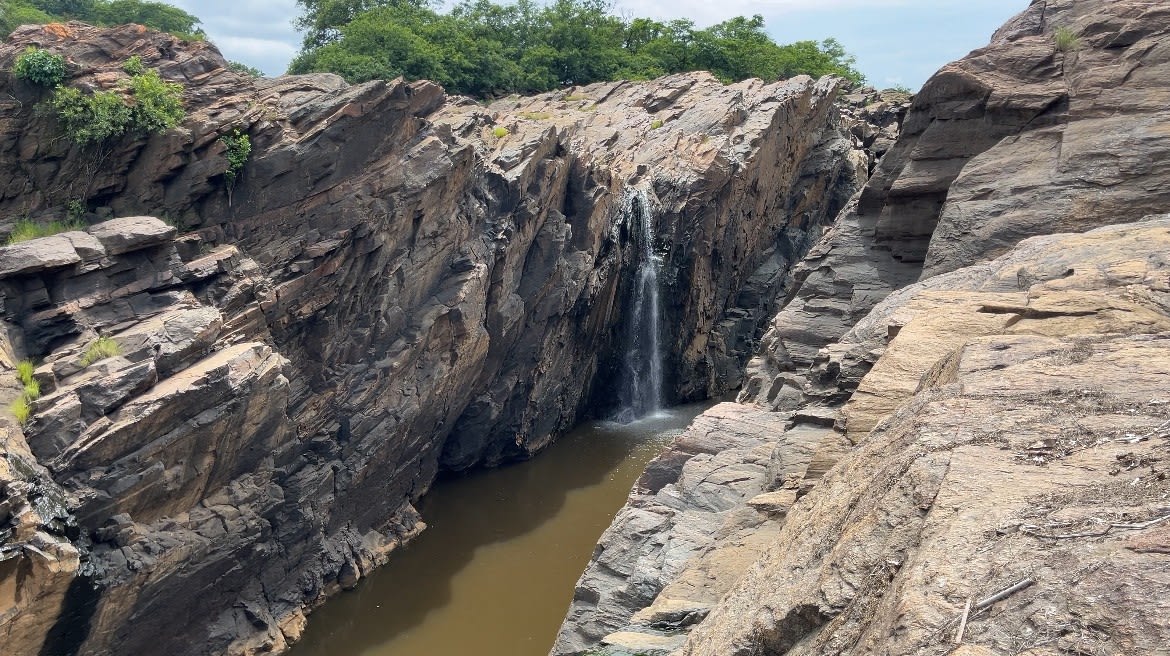 a narrow canyon with a small waterfall cascading down a steep, rugged, and jagged rock face into a calm channel of muddy brown water at the bottom.