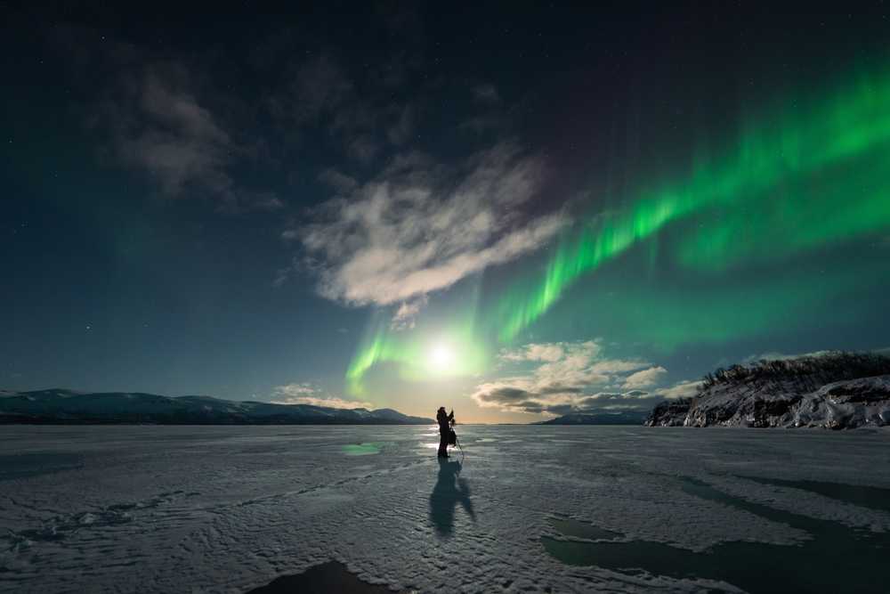 Northern lights above a frozen Iceland lake with a photographer under the night sky