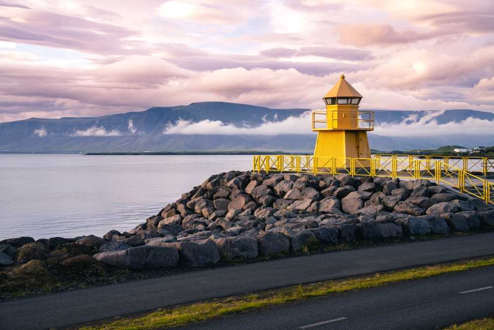 Yellow lighthouse on a rocky shoreline near Reykjavík with calm water and cloudy skies
