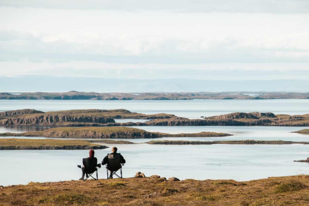 Two travelers sitting in camping chairs overlooking quiet Icelandic islands and water.
