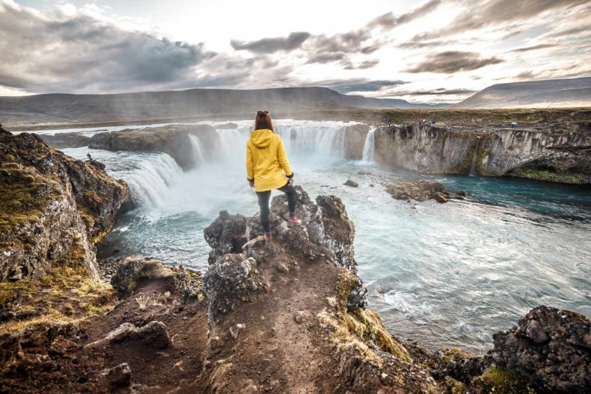Reisender in einer gelben Jacke mit Blick auf den Goðafoss-Wasserfall in Nordisland.