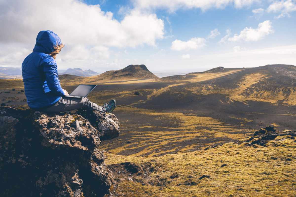 Reisende/r, der/die einen Laptop auf einem Felsen benutzt und dabei eine weite isländische Landschaft überblickt.