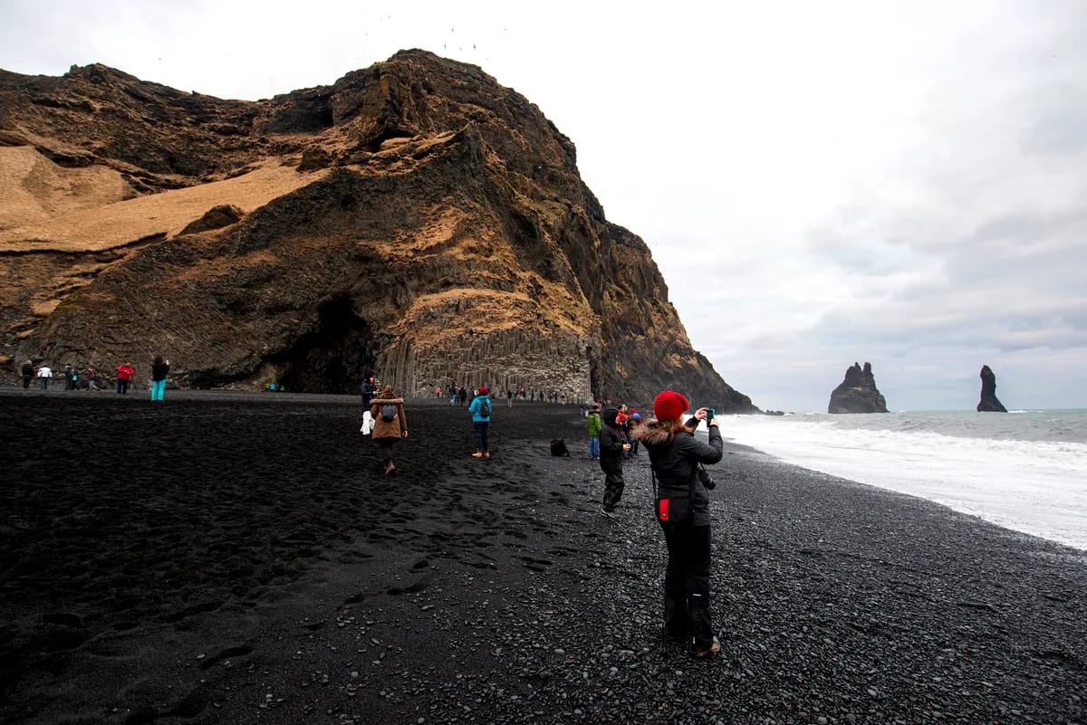 Tourists exploring Reynisfjara black sand beach, basalt cliffs and Reynisdrangar sea stacks under cloudy March skies in South Iceland
