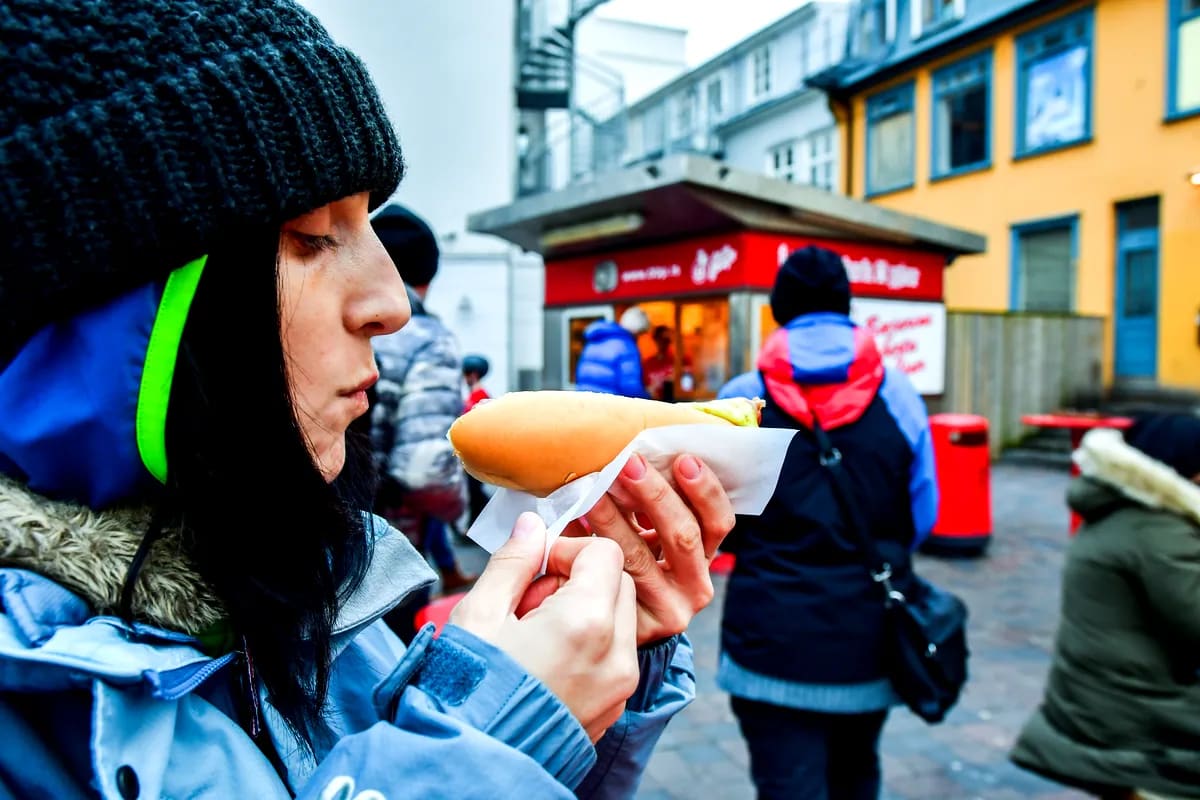 Traveler eating an Icelandic hot dog in Reykjavík, bundled in winter clothing beside a street food stand and colorful city buildings