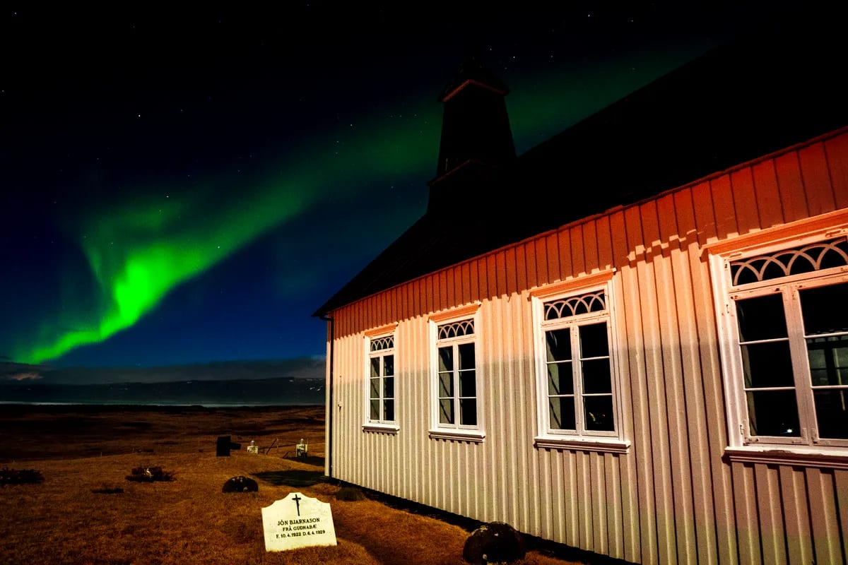 Northern Lights glowing over a white Icelandic church and graveyard at night, with green aurora, starry sky, and rural March landscape