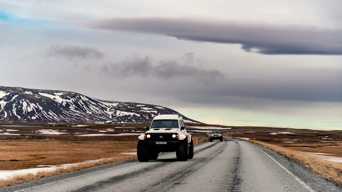 Super Jeep driving along an Iceland road in March, with snowy mountains, brown winter plains, wet pavement, and dramatic cloudy skies