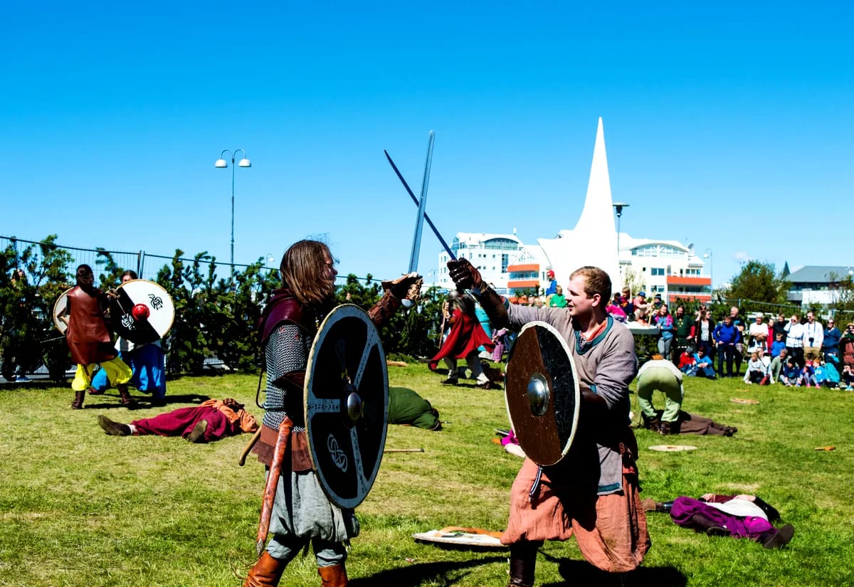 Viking reenactors with shields and swords performing in Reykjavík, with spectators on a green lawn and Hallgrímskirkja under bright Iceland skies
