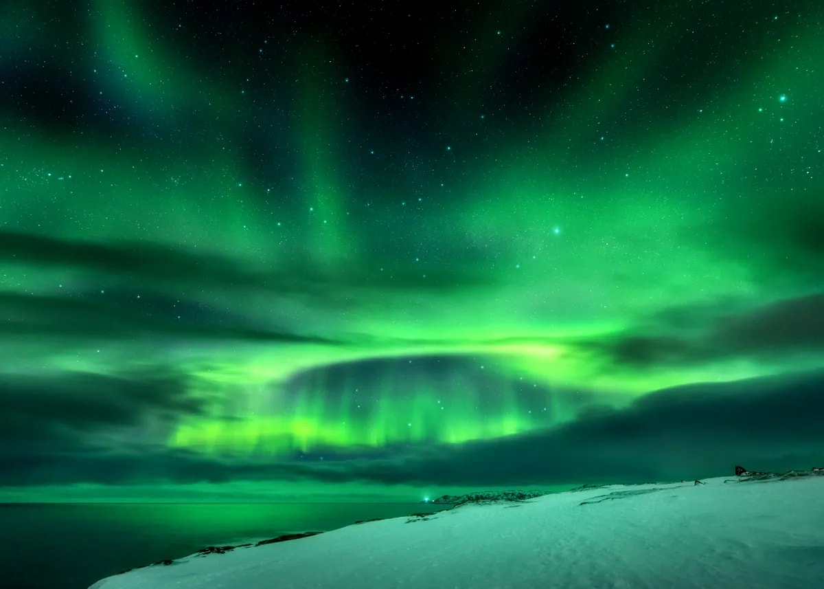 Bright green Northern Lights glow across a starry Iceland sky above snowy coastal hills, dark clouds, and a calm frozen seascape.