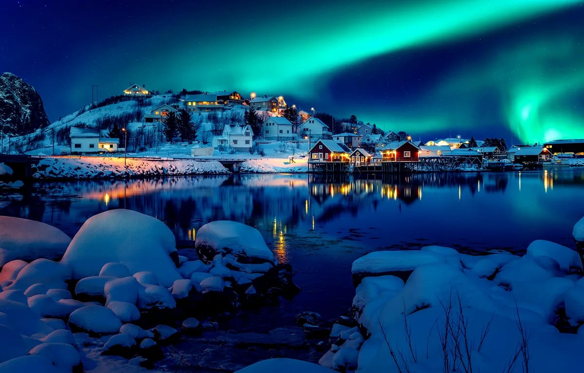 Green Northern Lights sweep over a snowy Icelandic harbor village, with warm cabin lights reflecting on calm water beneath a starry sky.