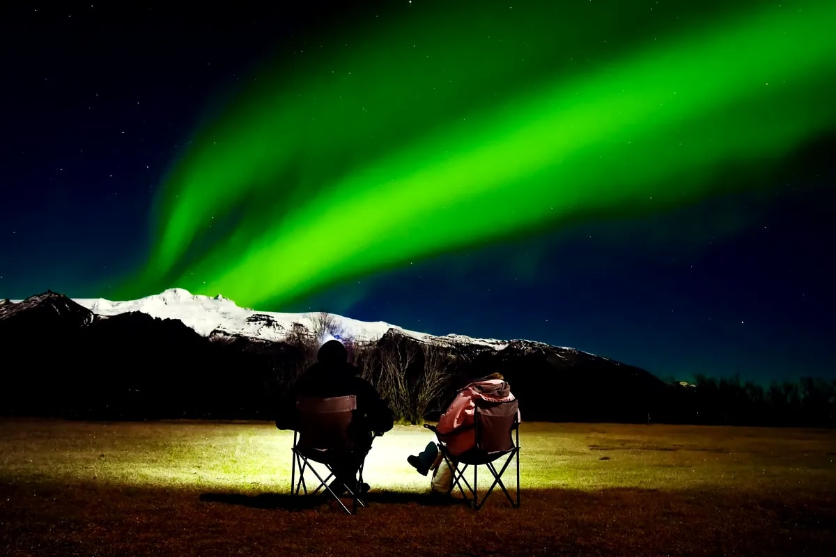 Two travelers sit in camp chairs watching vivid green Northern Lights sweep over snowy Iceland mountains under a clear starry night sky.