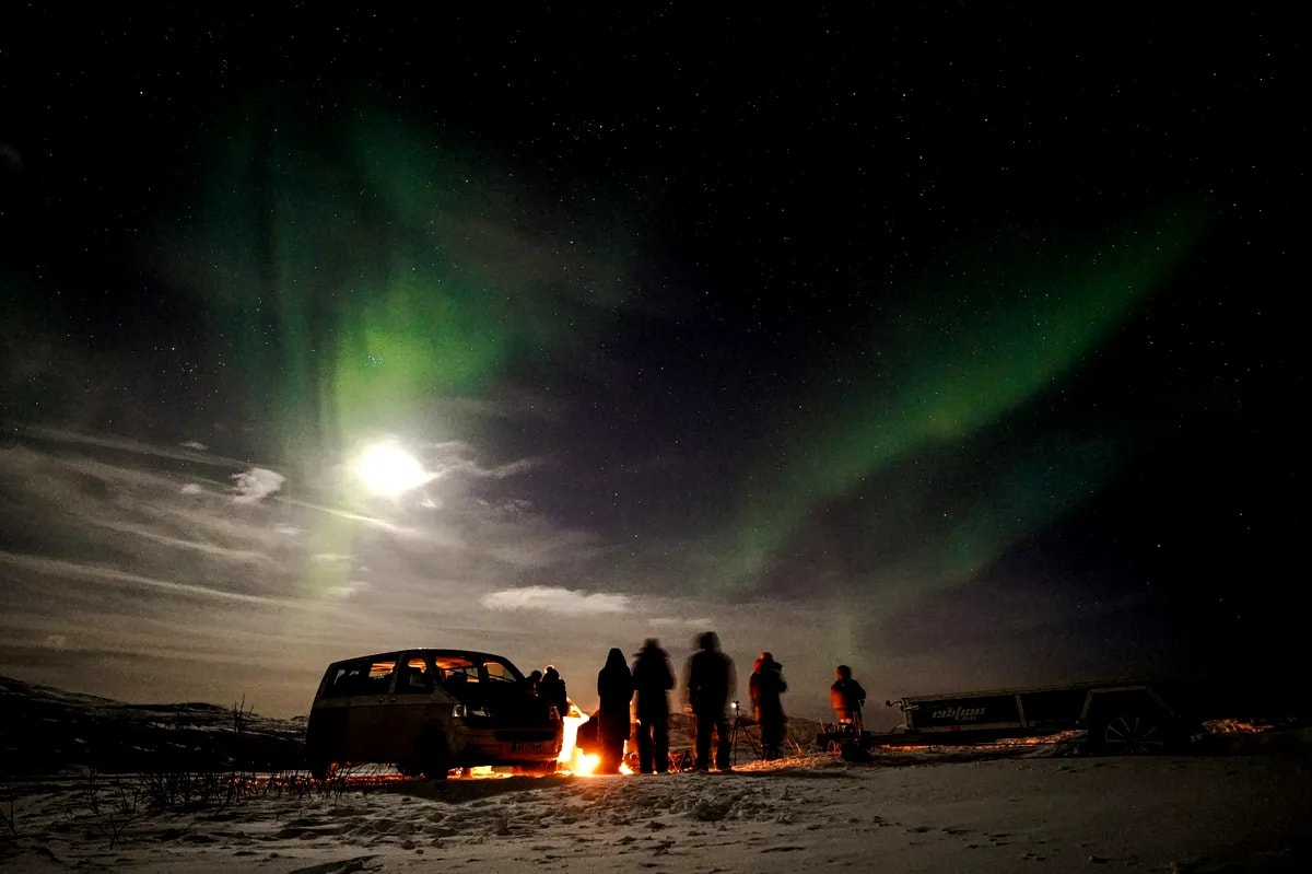 Silhouetted travelers gather by a campfire beside a van on snowy Iceland plains, with green Northern Lights, full moon, and stars.