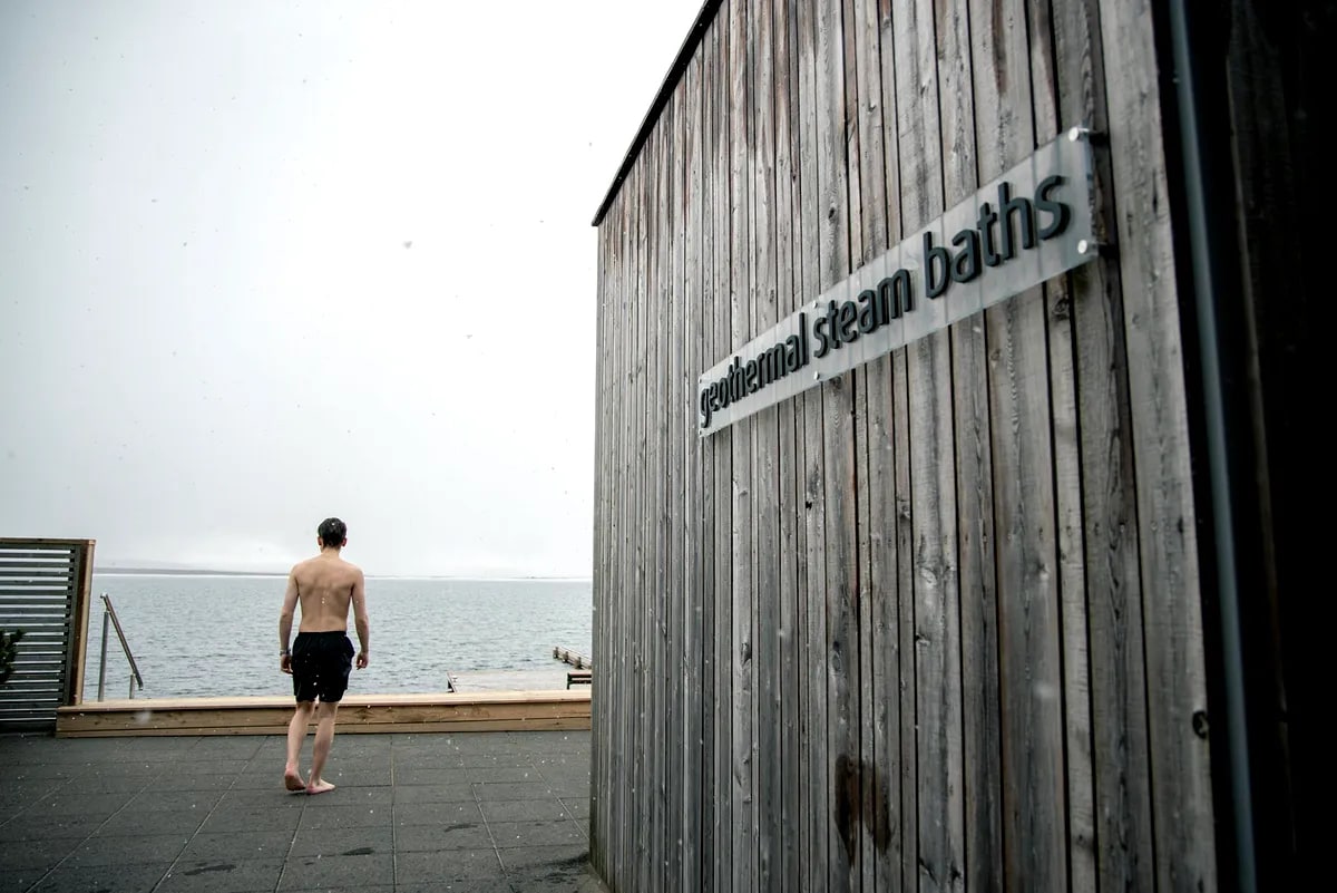 Visitor walking barefoot toward Laugarvatn Fontana lake beside wooden geothermal steam baths building on a misty Iceland spa day