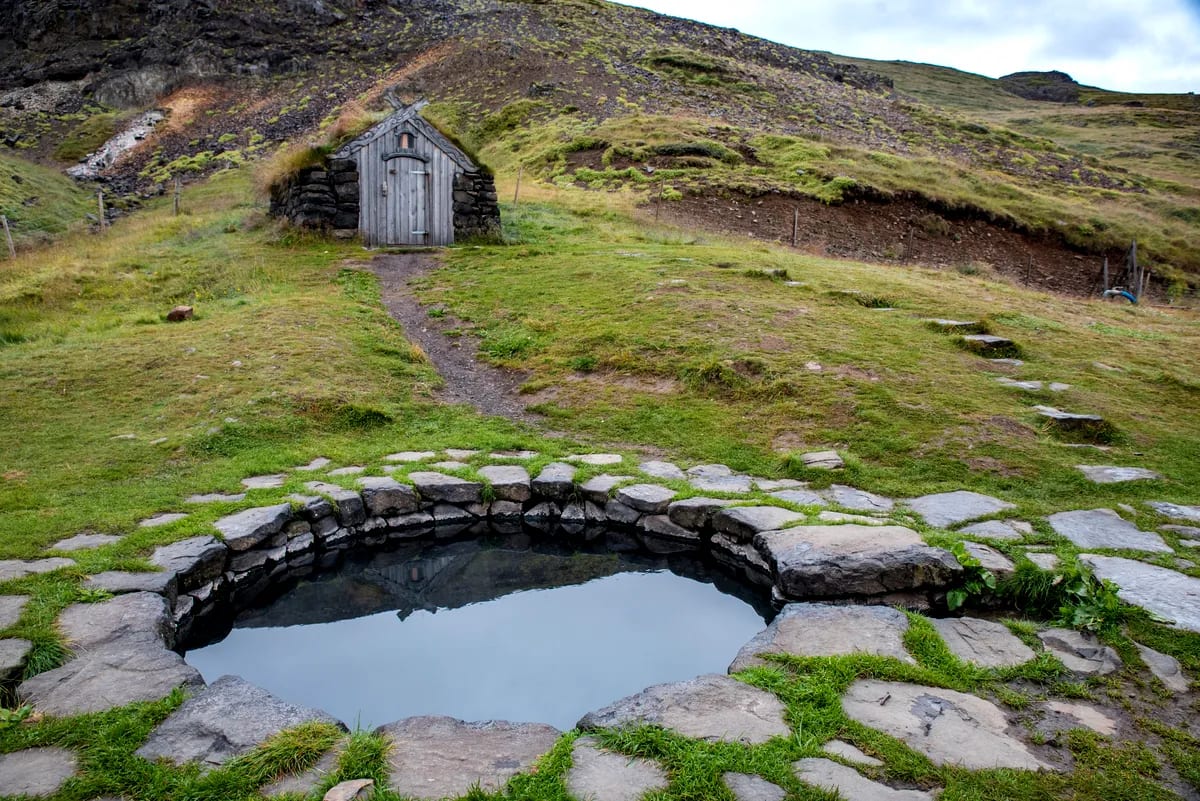 Guðrúnarlaug hot spring stone pool reflecting a turf-roof hut, surrounded by mossy hills and rugged Icelandic highland scenery