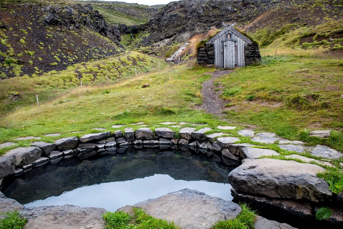 Guðrúnarlaug hot spring stone pool beside a turf-roof hut, framed by mossy cliffs, rocky gorge and quiet Icelandic valley trail