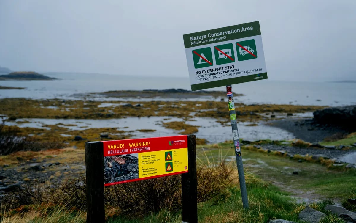 Warning and nature conservation signs at Hellulaug Hot Spring in Iceland, overlooking a misty rocky coast and quiet seaside landscape