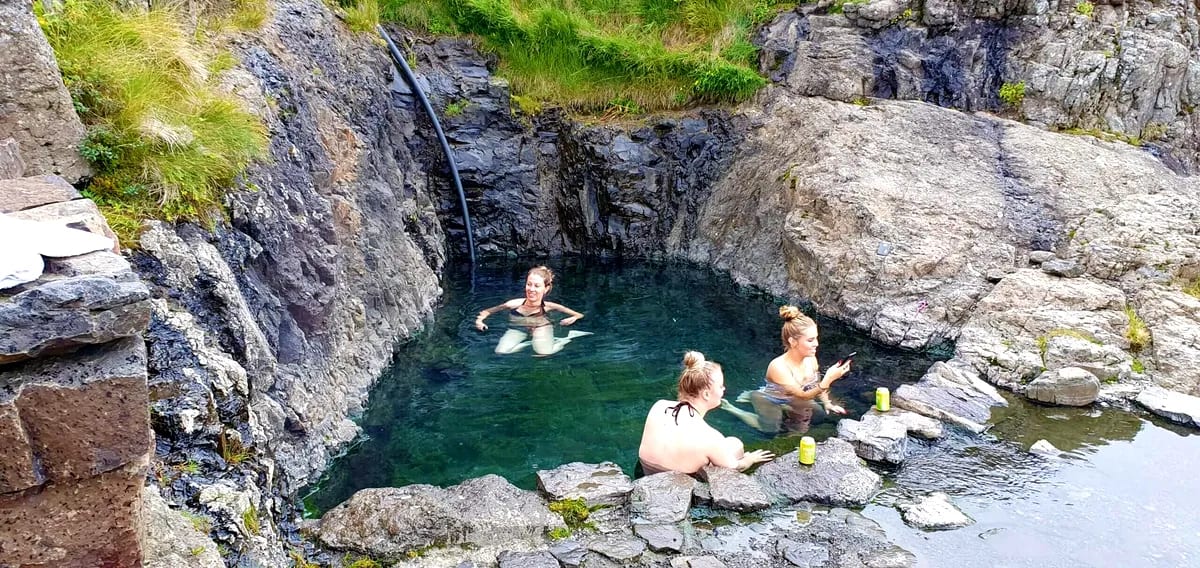 Visitors relaxing in Hellulaug Hot Spring’s clear geothermal pool, surrounded by rugged rocks and green grass on Iceland’s coast