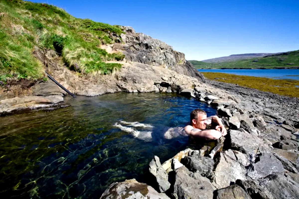 Man relaxing in Hellulaug Hot Spring's rocky geothermal pool with clear water, green cliffs, blue sky and coastal fjord views in Iceland