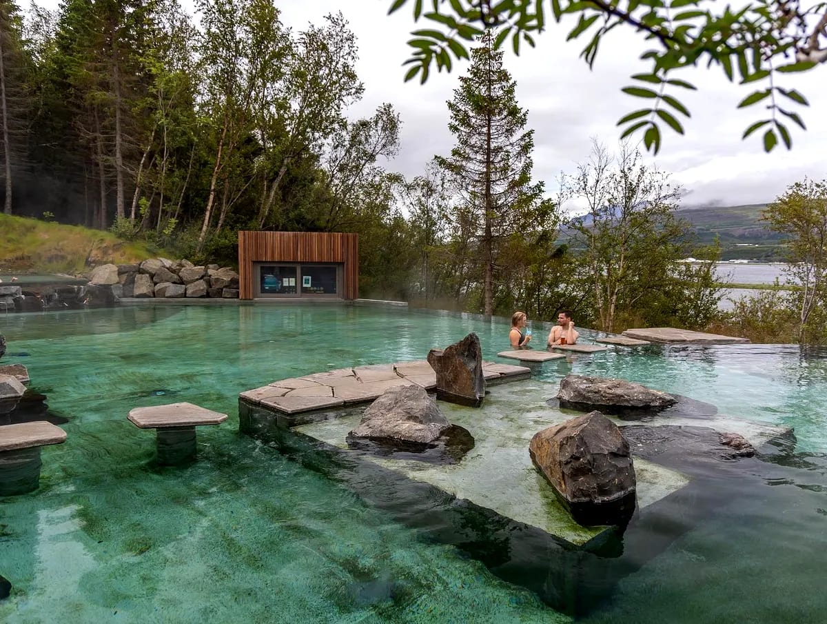 Couple relaxing in Forest Lagoon’s clear geothermal pool in Iceland, with volcanic rocks, birch forest, fjord views and misty mountains.
