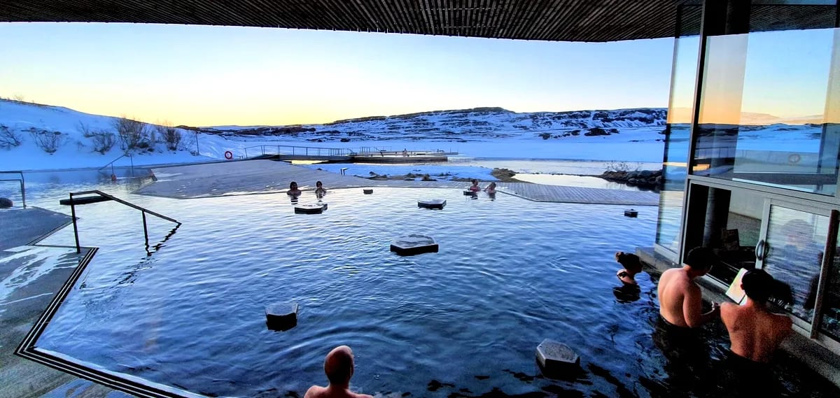 Guests soak in the warm outdoor geothermal pool at Vok Baths in Iceland, overlooking snowy hills and a frozen lakeside winter landscape