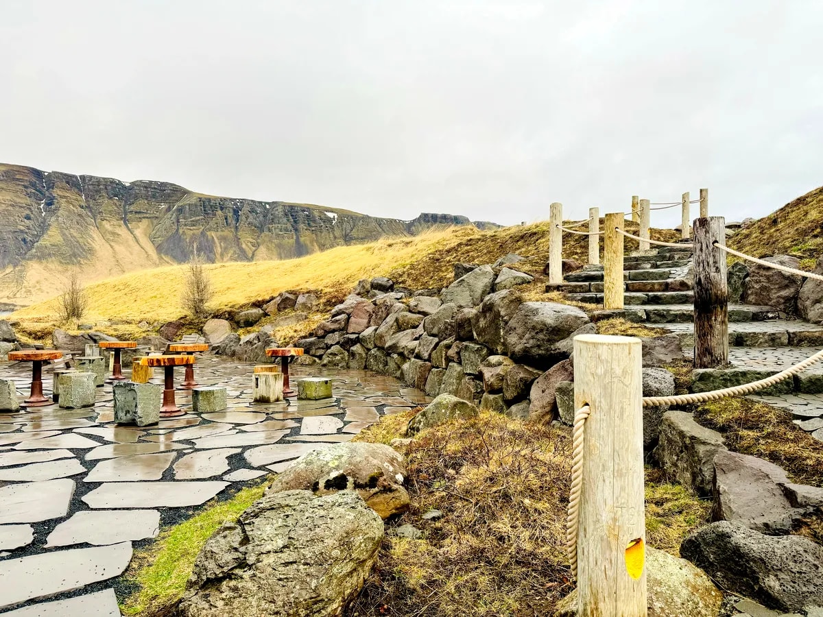 Stone terrace and rope-lined stairs at Hvammsvík Hot Springs, with volcanic rocks, wooden tables and dramatic Icelandic mountains